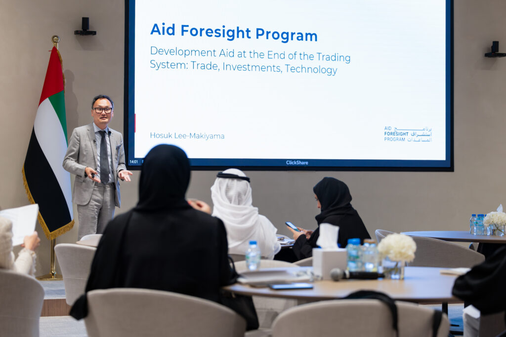 Hosuk Lee-Makiyama stands between a UAE flag, on the left of the screen, and a video screen that reads "Aid Foresight Program - Development Aid at the End of the Trading System: Trade, Investments, Technology"